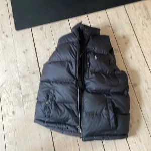 Ralph lauren dunväst med dragkedja - Säljer en svart dunväst från Ralph Lauren med hög krage och dragkedja framtill. Västen har två stora fickor med dragkedja och en mindre bröstficka. Perfekt för lager-på-lager och håller dig varm under kyliga dagar. Man kan även vända västen ut och in. Priset är 350kr men kan sänkas storlek 10-12 år 