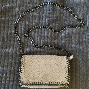 Beige axelväska med kedja och ormskinnsmönster - Säljer en beige axelväska med snyggt ormskinnsmönster och kraftig silverfärgad kedja som axelrem. Väskan har dekorativa kedjedetaljer runt kanterna och är perfekt för dig som vill ha en cool accessoar med lite edge.