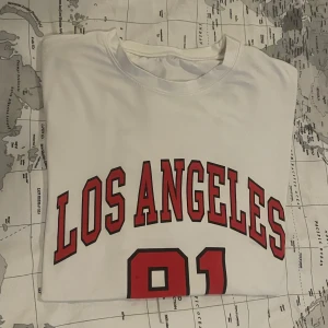Vit t-shirt med Los Angeles-tryck - Vit t-shirt med stort rött och svart tryck där det står 'LOS ANGELES 91' på framsidan. Klassisk rund halsringning och korta ärmar. 