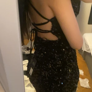 Svart glittrig balklänning  - Supersnygg svart balklänning täckt av glittriga paljetter. Klänningen har smala axelband som korsas i ryggen, öppen rygg med snörning och en hög slits på ena sidan. Perfekt för dig som vill sticka ut på festen!