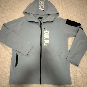 Ljusblå kofta från Hoodrich - Säljer en ljusblå hoodiejacka från Hoodrich med vit logga framtill och tryck på ryggen. Jackan har huva, dragkedja och svarta detaljer vid fickor och axel. Perfekt för en streetwear-look.