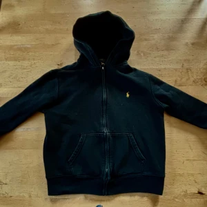 Svart hoodie från Polo Ralph Lauren - Klassisk svart hoodie från Polo Ralph Lauren med dragkedja framtill och huva. Tröjan har fickor på magen och den ikoniska lilla gula loggan broderad på bröstet. Perfekt för en avslappnad och stilren look. Priset kan diskuteras vid snabb affär 🤝
