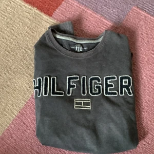 Mörkgrå sweatshirt från Tommy Hilfiger - Säljer en mörkgrå sweatshirt från Tommy Hilfiger med stor broderad logga framtill. Tröjan har rund halsringning och långa ärmar. Perfekt för en avslappnad och stilren look.