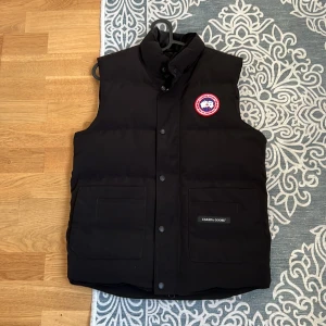 Svart dunväst från Canada Goose - Säljer en svart dunväst från Canada Goose med klassisk logotyp på bröstet och två stora fickor framtill. Västen har hög krage, knappar och dragkedja. Perfekt för kyliga dagar.