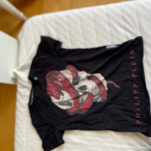 Svart t-shirt med ormtryck från Philipp Plein - Cool svart t-shirt från Philipp Plein med stort tryck av en röd och vit orm på framsidan. Märkets namn står tryckt längs sidan i rött. Klassisk passform och korta ärmar, perfekt för dig som vill sticka ut.