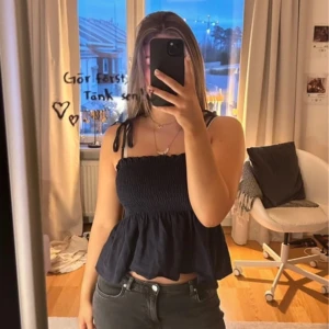 Svart smockad linnetopp med knytband - Säljer en svart linnetopp med smockad överdel och volang nertill. Toppen har smala axelband med knyt på axlarna och en luftig passform. Perfekt för varma dagar eller att styla lager på lager.