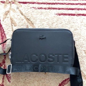 Svart axelväska från Lacoste - Säljer en svart axelväska från Lacoste med tydlig logotyp framtill och justerbar axelrem. Väskan har dragkedja upptill och är tillverkad i ett slitstarkt syntetmaterial. 