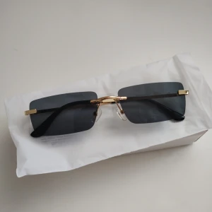 Svarta rektangulära solglasögon med gulddetaljer - Stilrena solglasögon med rektangulära svarta glas och tunna guldfärgade bågar. Glasögonen har näskuddar och svarta skalmar. Perfekta för dig som vill ha en modern och minimalistisk look.