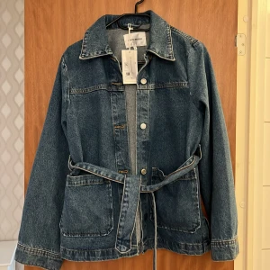Jeansjacka med bälte från Carin Wester - Säljer en klassisk blå jeansjacka från Carin Wester med bälte i midjan och stora fickor framtill. Jackan har långa ärmar, knappar och en avslappnad passform. Perfekt för lager-på-lager-stil.