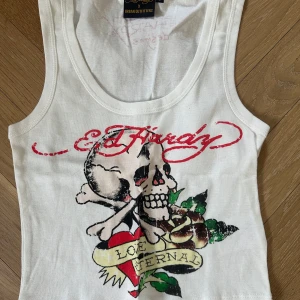 Vit linnetopp från Ed Hardy x Urban Outfitters - Säljer en vit linnetopp från Ed Hardy x Urban Outfitters med tryck av dödskalle, ros och texten 'Love Eternal' framtill. Toppen har ärmlös design och rund halsringning. Pris kan diskuteras 
