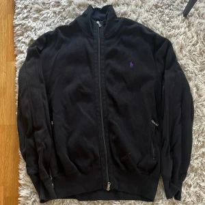 Svart stickad full zip från Ralph Lauren - Svart stickad full zip  med dragkedja från Ralph Lauren. Tröjan har hög krage, två fickor med dragkedja och en liten lila broderad logga på bröstet. Perfekt för en avslappnad stil.