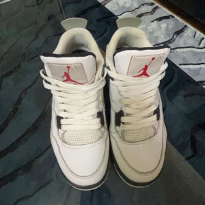 Nike Air Jordan 4 - Säljer ett par vita Nike Air Jordan 4 detaljer och svarta inslag. Skorna har klassisk mesh på tån, synlig snörning och specklad grå häl med Nike Air-logga. Perfekta för dig som gillar streetwear och sneakerskultur.