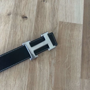 Svart skärp med silverfärgat H-spänne - Säljer ett svart skärp från H med ett stort H-format spänne i silverfärg. Skärpet är gjort i skinn med vit kontrastsöm längs kanterna och har flera hål för justering. Perfekt accessoar för att lyfta din outfit.