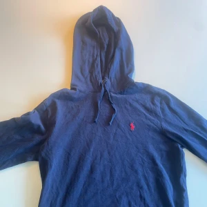 Marinblå hoodie från Polo Ralph Lauren - Säljer en marinblå hoodie från Polo Ralph Lauren med klassisk röd broderad logga på bröstet. Tröjan har huva med dragsko och långa ärmar. Perfekt för en avslappnad och stilren look.