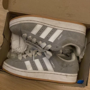 Grå Adidas Campus sneakers - Säljer ett par grå Adidas Campus sneakers med vita ränder och vit sula. Skorna har klassisk låg modell, breda vita skosnören och är tillverkade i mocka. Perfekta för en stilren vardagslook.
