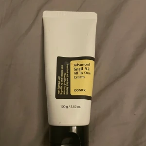 COSRX Advanced Snail 92 All In One Cream - En ansiktskräm med 92% snigelsekretfiltrat som hjälper till att återfukta och stärka huden. Kommer i en vit tub med svart och gul etikett, innehåller 100 g. Perfekt för dig som vill ha glow och mjuk hud. Andvänd