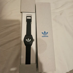 Svart klocka från Adidas - Säljer en stilren svart klocka från Adidas med klassisk logga på urtavlan. Klockan har ett svart armband och levereras i originalförpackning med Adidas-tryck. Perfekt accessoar för dig som gillar sportig och enkel design.