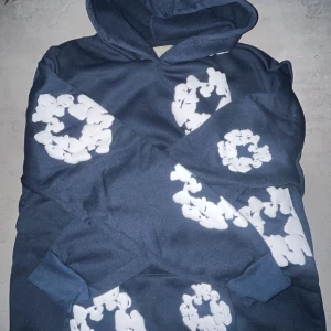 Mörkblå hoodie med vita blommönster från Denim tears - Säljer en mörkblå hoodie från Denim tears med stora vita blommönster tryckta över hela tröjan. Tröjan har huva och magficka, perfekt för en avslappnad stil.