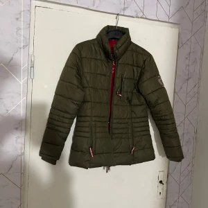 Olivgrön quiltad jacka från Covalliero - Säljer en olivgrön quiltad jacka från Covalliero med röd insida och broderad logga på ärmen. Jackan har dragkedja, hög krage och flera fickor med dragkedja. Perfekt för dig som gillar ridning och vill ha en snygg och funktionell jacka.