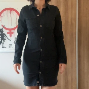 Svart jeansklänning med knappar - Säljer en svart jeansklänning med lång ärm och knappstängning hela vägen framtill. Klänningen har krage och en figurnära passform som ger en snygg siluett. Perfekt för dig som gillar en stilren och enkel look.