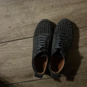 Svarta sneakers med nitar och röd sula - Säljer ett par svarta sneakers med coola nitar på tån och klassisk röd sula. Skorna har snörning och en platt sula. Perfekta för dig som vill sticka ut med din stil.