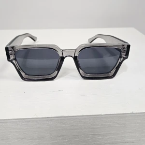 Grå fyrkantiga solglasögon - Snygga solglasögon med markerad, transparent grå båge och mörka glas. Glasögonen har en modern, kantig form som ger en cool och trendig look. Perfekta för dig som vill sticka ut med accessoarer i vardagen.Snygga solglasögon med markerad, transparent grå båge och mörka glas. Glasögonen har en modern, kantig form som ger en cool och trendig look. Perfekta för dig som vill sticka ut med accessoarer i vardagen.