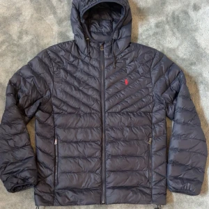 Ralph Lauren jacka  - Säljer en svart pufferjacka från Polo Ralph Lauren med huva och dragkedja framtill. Jackan har två fickor med dragkedja och en liten röd logga på bröstet. Perfekt för kyliga dagar.