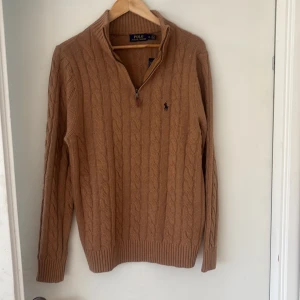 Beige kabelstickad tröja från Polo Ralph Lauren - Säljer en beige kabelstickad tröja från Polo Ralph Lauren med halv dragkedja och broderad logga på bröstet. Tröjan har lång ärm och ribbade muddar. Perfekt för lager-på-lager och snygg till både jeans och chinos. Jag är 189cm - 89kg