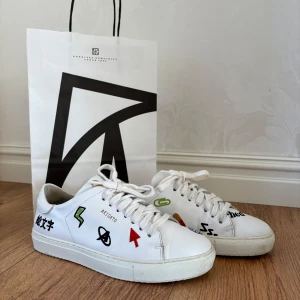 Vita sneakers med färgglada patches från Axel Arigato - Säljer ett par vita sneakers från Axel Arigato med coola färgglada patches och broderade detaljer. Skorna har snörning och platt sula, samt rund tå. Perfekta för dig som vill sticka ut med din stil!