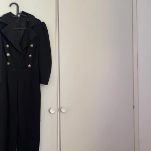 Svart jumpsuit med guldknappar - Snygg svart jumpsuit med markerade axlar och dubbelknäppt design framtill. Guldiga knappar ger en lyxig känsla och den har långa ben samt djup v-ringning. Perfekt för dig som vill sticka ut.