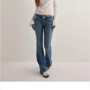 Blå bootcut jeans från True Religion - Säljer ett par klassiska blå bootcut jeans från True Religion med ikoniska bakfickor och rosa sömmar. Jeansen har låg midja och femficksdesign, perfekt för en avslappnad och trendig look.