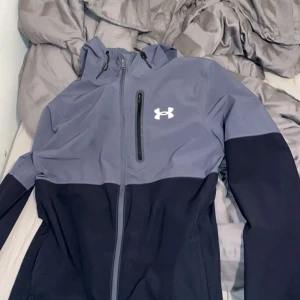 Under Armour tvåfärgad träningsjacka med huva - Snygg träningsjacka från Under Armour i grått och svart. Jackan har huva, dragkedja framtill och en bröstficka med dragkedja. Perfekt för träning eller aktiva dagar. Under Armour-logga på bröstet.