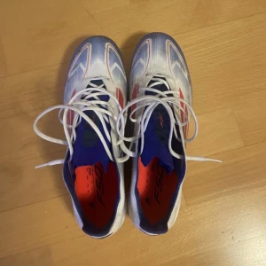 Adidas f50 elite - Säljer ett par Adidas fotbollsskor i vitt med blå och röda detaljer. Skorna har snörning och mönstrad ovandel samt dobbar i olika färger under sulan. Perfekta för dig som vill sticka ut på planen!