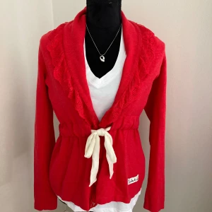 Cardigan  med spetsdetaljer från Bondelid - Cardigan från Bondelid med fina spetsdetaljer längs kragen och ett vitt knytband i midjan. Jackan har lång ärm och en  volangkant. 