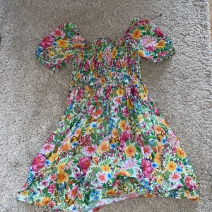 Blommig sommarklänning med puffärmar - Färgglad blommig klänning i ekologisk bomull med smock upptill och puffiga korta ärmar. Klänningen har en vid kjol och massor av fina blommor i rosa, gul, orange och grönt. Perfekt för varma dagar och ger en riktigt somrig känsla.