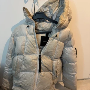 Beige dunjacka från Peak Performance - Säljer en beige dunjacka från Peak Performance med huva och dragkedja framtill. Jackan är quiltad och har en sportig look, perfekt för kyliga dagar. Märkeslogga på ärmen och praktiska fickor. Snygg och funktionell jacka för vintern.