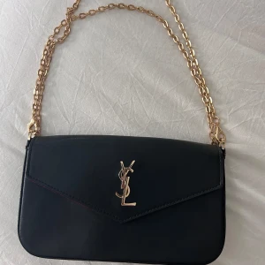 Svart axelväska från Saint Laurent - Säljer en svart axelväska från Saint Laurent med guldfärgad kedja och logga framtill. Väskan har ett rött innerfoder och magnetlås. Perfekt för att lyfta vilken outfit som helst.