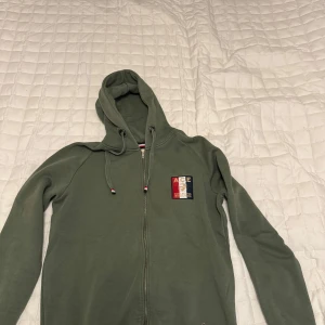 Grön hoodie med dragkedja från A.C.E Collective Evolution - Säljer en grön hoodie från A.C.E Collective Evolution med dragkedja och huva. Tröjan har ett broderat märke på bröstet i rött, vitt och blått samt mjukt foder på insidan. Perfekt för en avslappnad stil.