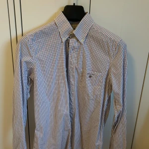 Gant Skjorta - Säljer en klassisk rutig skjorta från GANT i vitt med blå och röda rutor. Skjortan har lång ärm, knappar framtill och en bröstficka. Perfekt för dig som gillar stilrena och tidlösa plagg.
