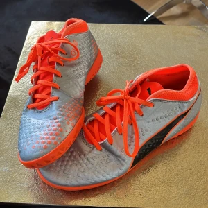 Silvriga och orangea fotbollsskor från Puma - Säljer ett par snygga fotbollsskor från Puma i silver med orange detaljer och svart logga. Skorna har snörning och en platt, mönstrad sula som passar för spel på konstgräs eller inomhus. Perfekta för dig som vill sticka ut på planen! Strl 40,5