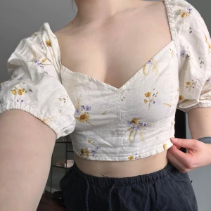 Vit croppad blus med blommigt mönster  - Säljer en söt croppad blus i vit bomull med blommigt mönster i gult och lila. Toppen har puffiga korta ärmar med resår och en v-ringad urringning. Perfekt för varma dagar och ger en romantisk känsla🎀