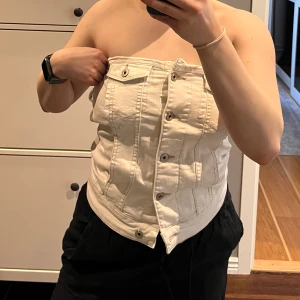 Vit jeans-korsett med knappar - Säljer en egengjord vit korsett-topp i jeanstyg med knappar framtill och dekorativa fickor. Gjorde den på en gammal jeansjacka! Toppen är axelbandslös och har en figurnära passform. Perfekt för att skapa en cool och edgy look. Tror den hade passat bättre en längre överkropp då den ”skrynklar” ihop på mig.