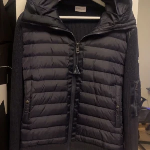 Moncler jacka - Säljer en svart Moncler-jacka med quiltad front och stickade ärmar. Jackan har huva, dragkedja framtill och fickor med tryckknappar. Klassisk Moncler-logga på ärmen och extra ficka med dragkedja på ena ärmen.