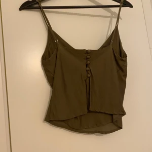 Olivgrön linnetopp med knappar - Säljer en olivgrön linnetopp med smala axelband och dekorativa knappar baktill. Toppen har en lätt asymmetrisk nederkant och är perfekt för varma dagar.