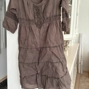 Brun tunn  kortklänning.tunika med spetsdetaljer från Vila - Säljer en brun tunn  kortklänning/tunika från Vila med fina spetsdetaljer vid halsringningen och midjan. Klänningen har trekvartsärm och ett avslappnat fall. Perfekt för dig som gillar bohemisk stil. Saknas resår runt halslinningen 