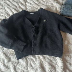 Svart kofta från Lacoste - Säljer en svart kofta från Lacoste med klassisk krokodillogga på bröstet. Koftan har knappar framtill och är i ett mjukt, stickat material. Perfekt för lager-på-lager och enkel att matcha med olika outfits.