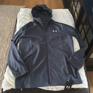 Mörkblå vindjacka från Under Armour - Säljer en mörkblå vindjacka från Under Armour med huva och dragkedja. Jackan har två sidofickor med dragkedja och logga på bröstet samt stor logga på ryggen. Perfekt för träning eller utomhusaktiviteter. Jackan är helt nya. Säljer den pågrund av att den inte passade mig den är köpt på jd sports