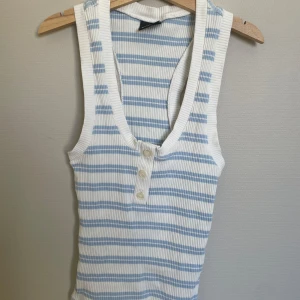 Randig ribbad linnetopp från Gina Tricot - Säljer en ribbad linnetopp från Gina Tricot i vitt och ljusblått med smala ränder. Toppen har djup v-ringning och knappar framtill. Perfekt för varma dagar och enkel att matcha med jeans eller shorts.