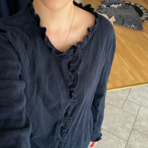 Jättefin marinblå kofta med räfflade ärmar - Mörkblå kofta med långa ärmar och knäppning framtill. Volangdetalj längs knappraden och halsringningen. Perfekt för lager-på-lager och enkel att matcha till olika outfits. 100% bomull.