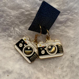 Kameraformade örhängen i svart och guld - Unika örhängen formade som små kameror i svart, vitt och guld. Perfekta för dig som älskar foto eller vill ha ett statement-smycke. Hängande modell med fina detaljer och retrovibbar. Krokarna är i sterling silver